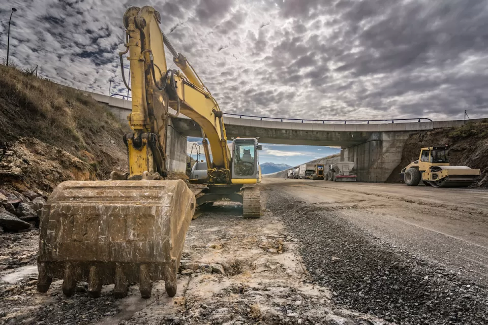 Excavator parked on side of highway under construction with overpass bridge in background.